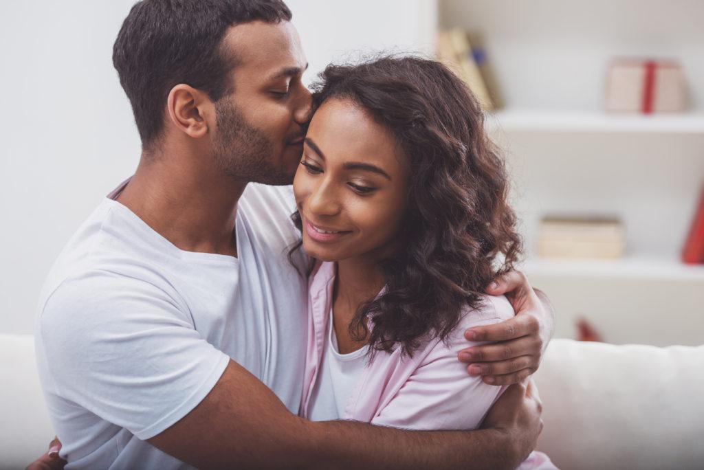 Young beautiful Afro-American couple hugging and smiling while sitting on a sofa in the room. Man kissing his woman.