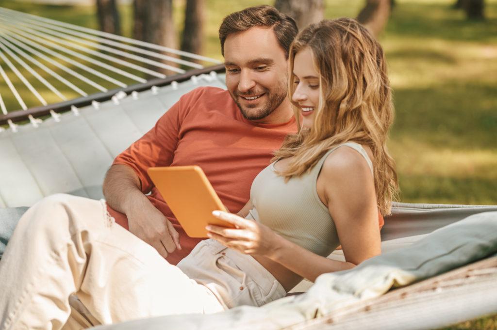 Good moments. Happy young smiling attractive man and charming woman with long blond hair looking interested in tablet on hammock outdoors